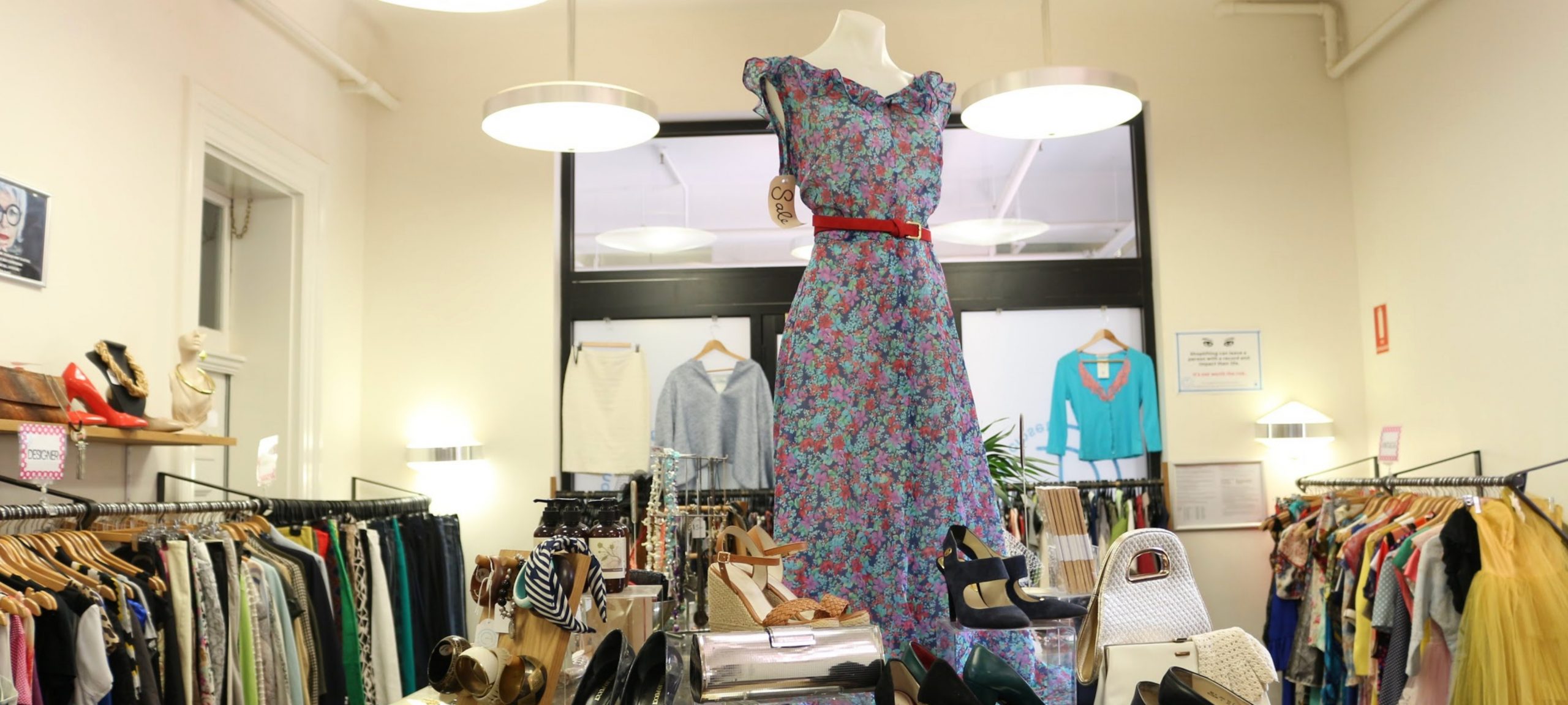 Masthead 3 The Conscious Closet store. A mannequin dressed in a purple floral dress sits on a table surrounded by stylish vintage clothing and accessories