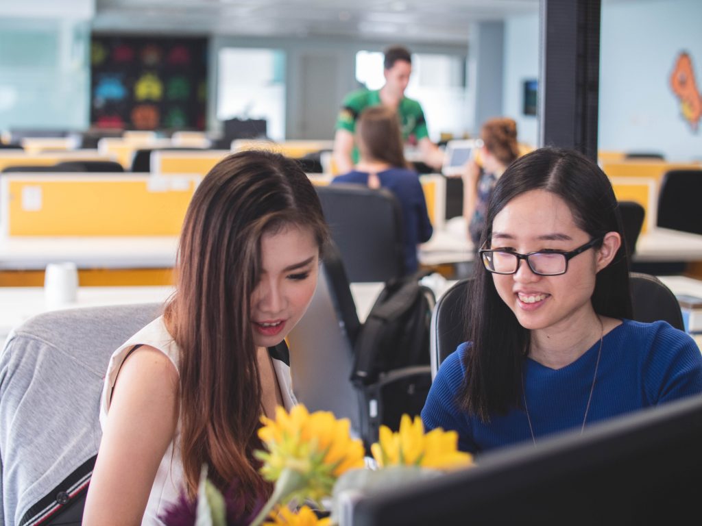 Two asian women sit at a computer working collaboratively. They are in a busy office with lots of shared working stations.