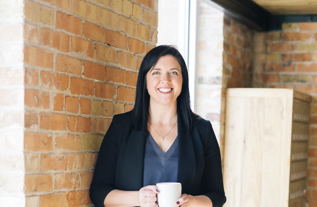 Tamara stands against a brick wall  holding a cup of coffee and smiling warmly. She has long brown hair and is wearing a stylish suit.