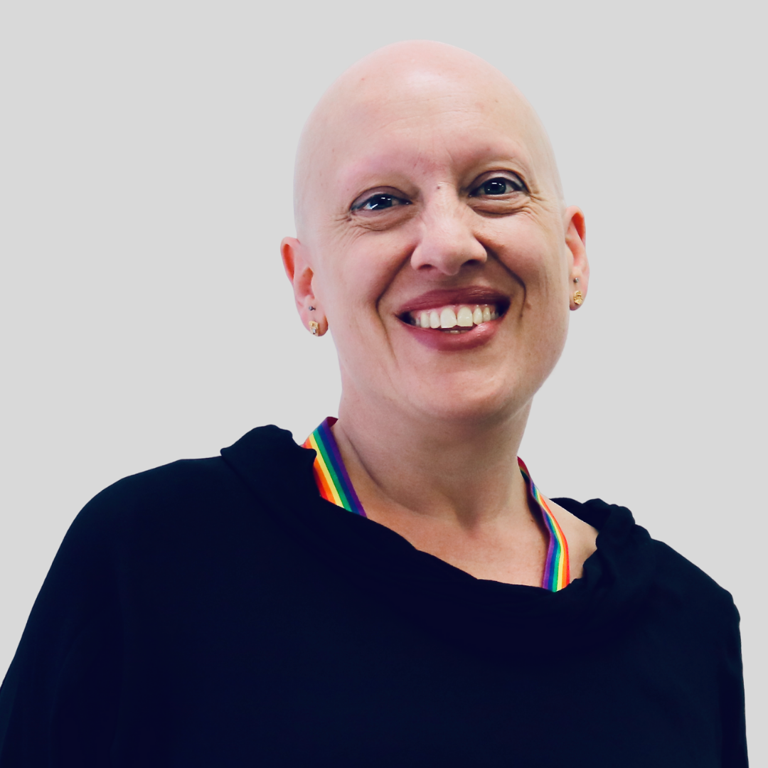 Headshot of Bek Pasqualini - bald woman in mid-life wearing rainbow lanyard