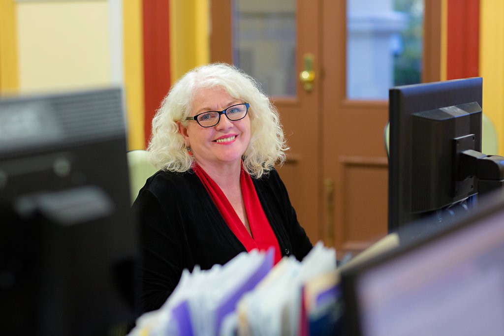 An older, professionally dressed woman sits at a work station, smiling.