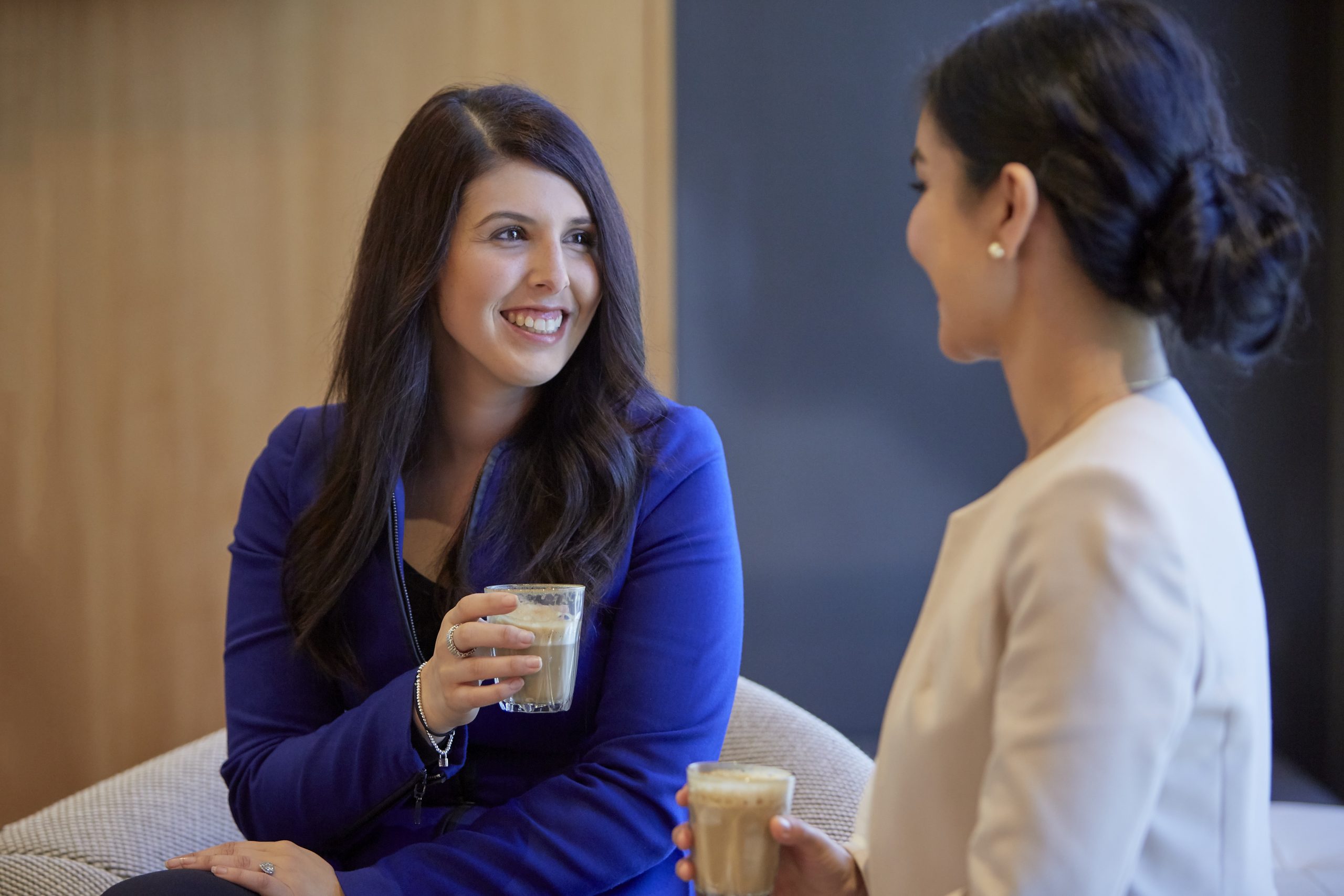 Two female corporate partners sit drinking a cup of coffee. The are wearing professional clothing and smiling