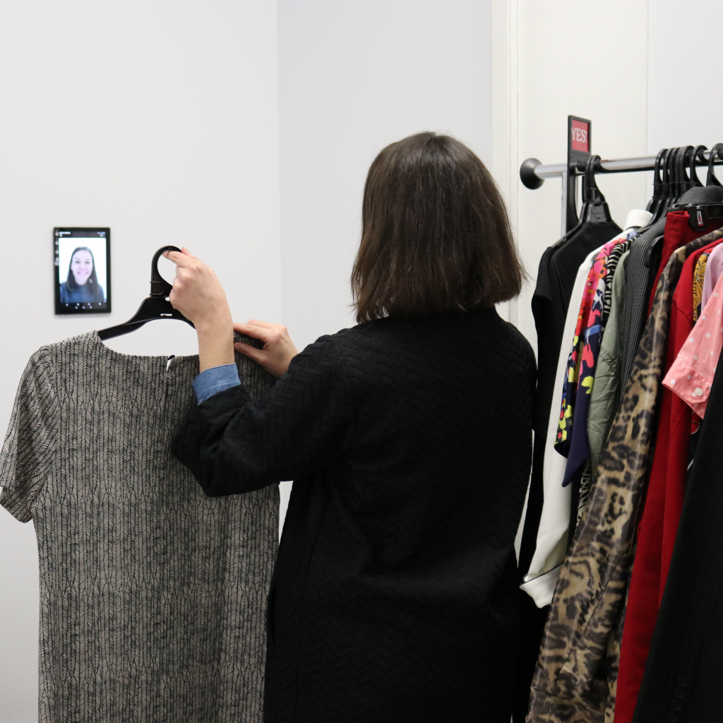 A woman with dark brown hair holds a professional dress up to video screen, where a woman is watching. Beside her is a rack of clothing.