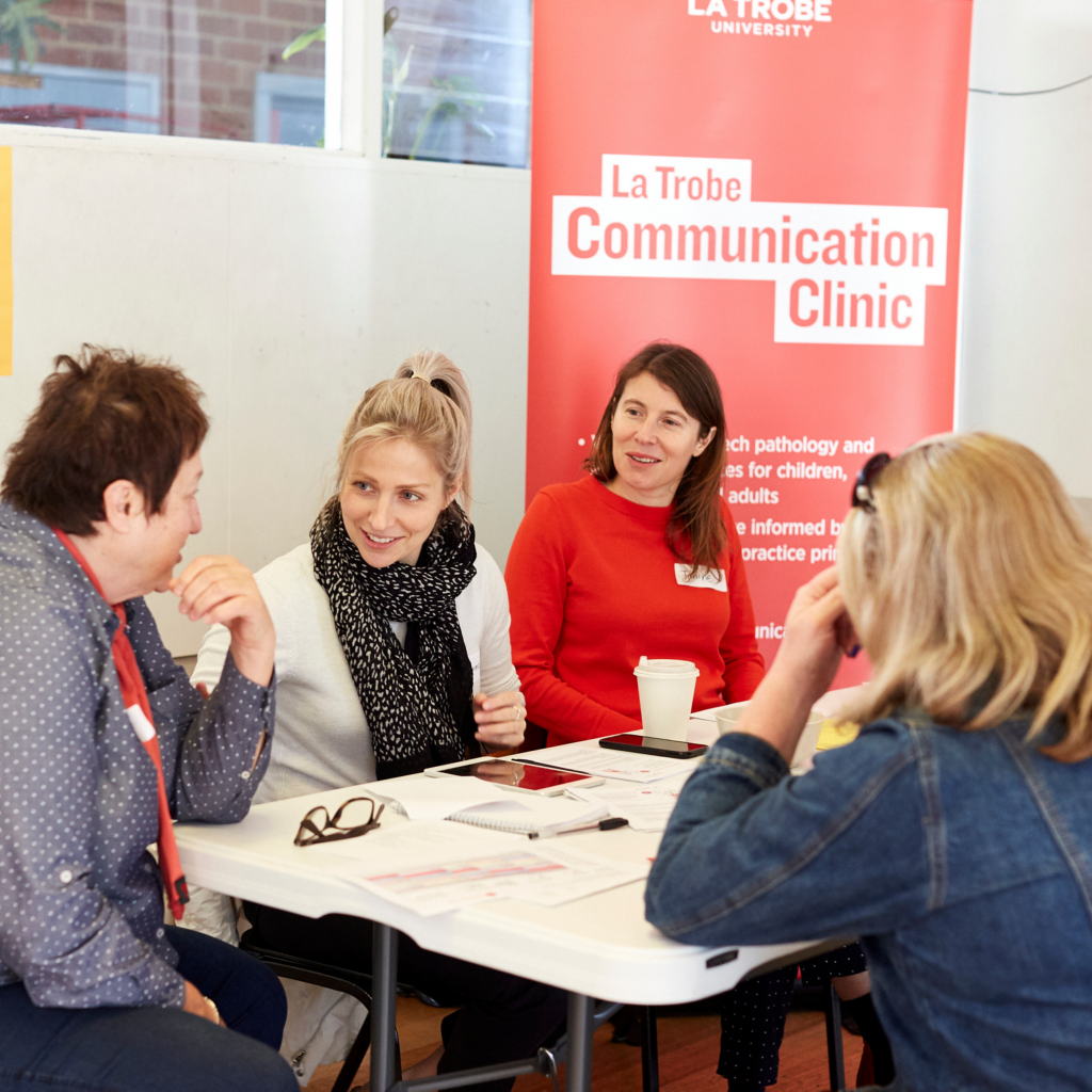 Two female volunteers sit at a table with two transgender women talking. Behind them is a red sign that says La Trobe Communication Clinic.