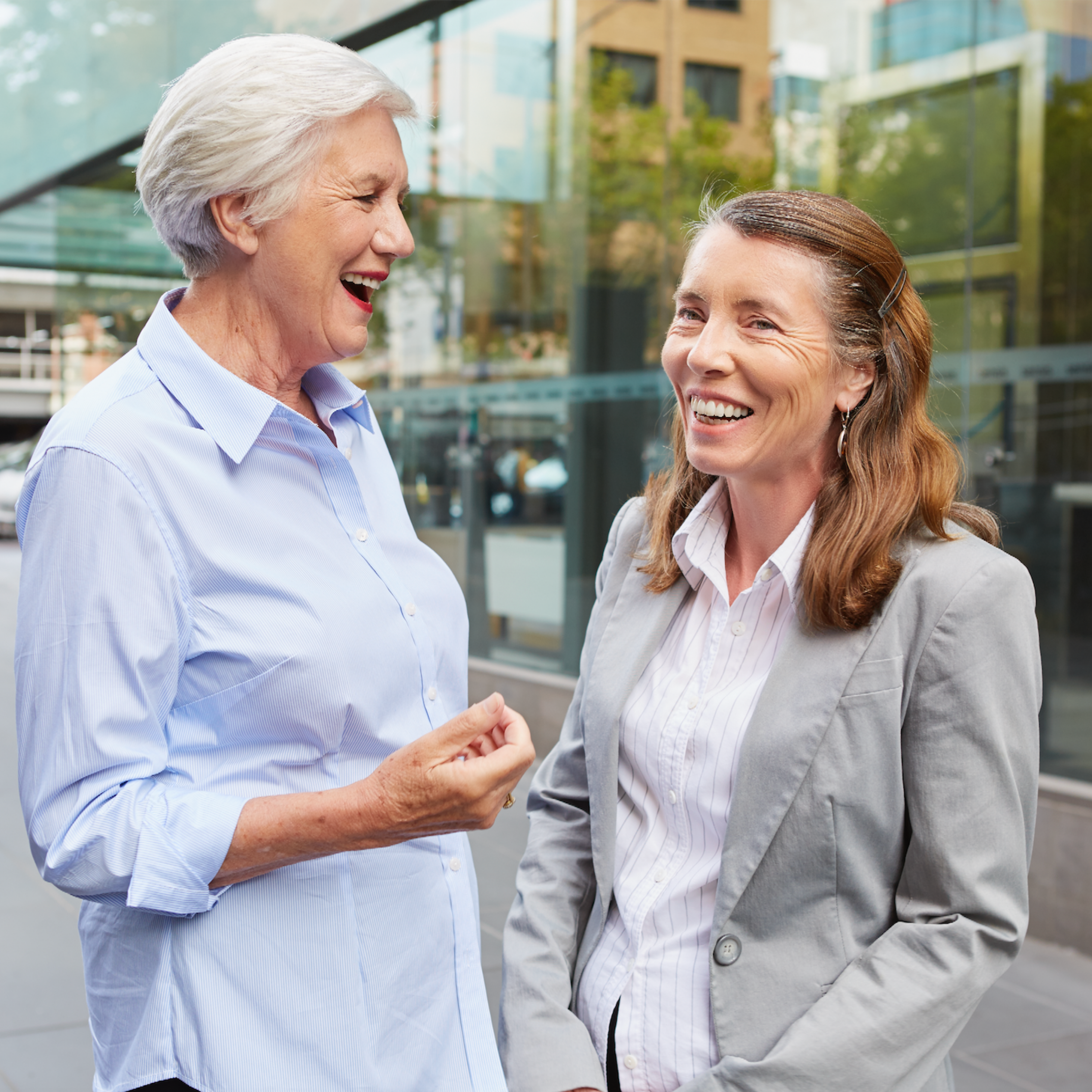 A Fitted for Work mentor and client stand on a street corner wearing professional clothing. They are facing each other and laughing.