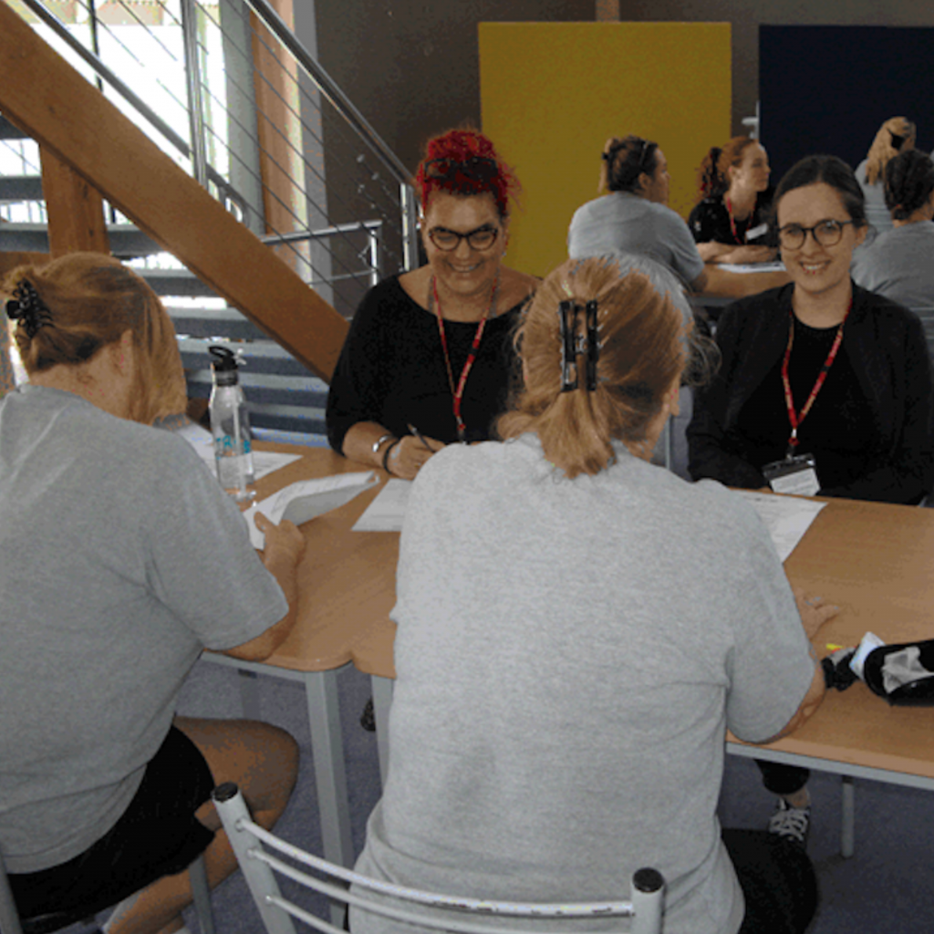 Image of two volunteers working with to women at a table. The two women are wearing grey shirts.