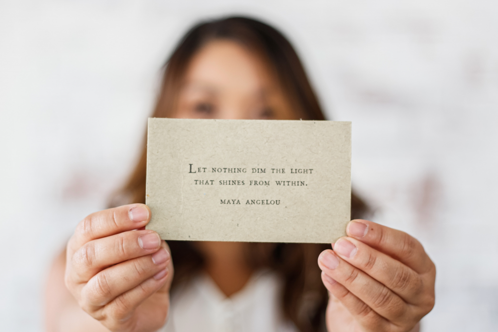 Woman with brown hair holds up a card that says 'let nothing dim the light that shines from within' Maya Angelou