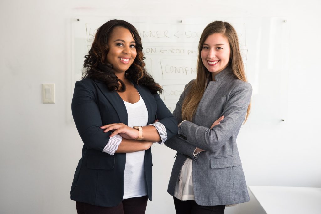 Two female corporate partners stand with their arms crossed. They are wearing business suits and smiling.