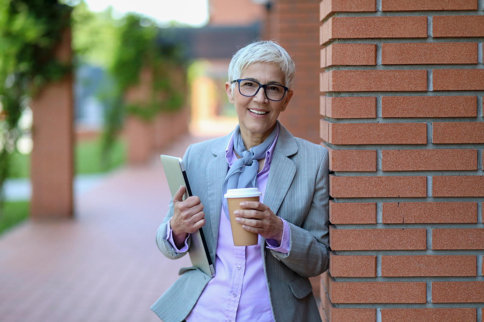 A woman with short grey hair and glasses, wearing a professional outfit is posing beside a red brick wall. She is holding a folder and a takeaway coffee and smiling warmly.