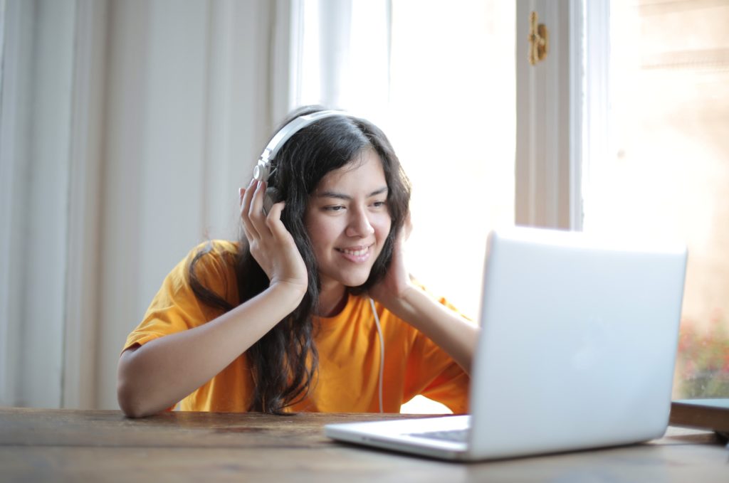 A young woman with dark hair is listening to a video on a laptop. She is wearing large headphones and holding them over her ears, listening intently.