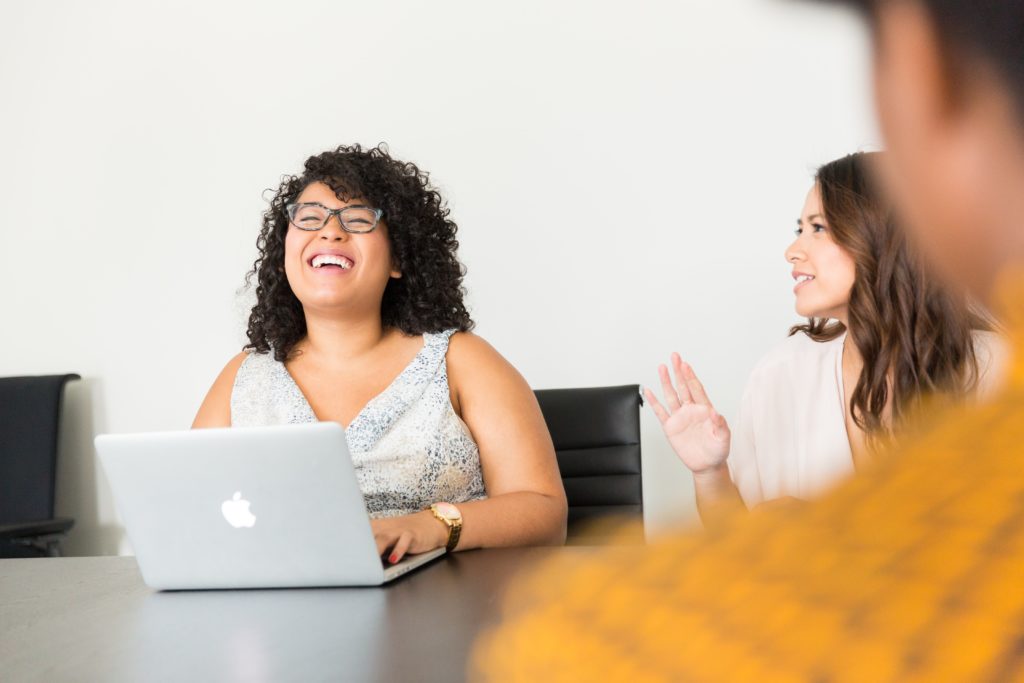 A woman with curly dark hair and glasses is typing on a computer, she is throwing her head back laughing as a brunette woman next to her talks animatedly. They are sitting in an office meeting room.