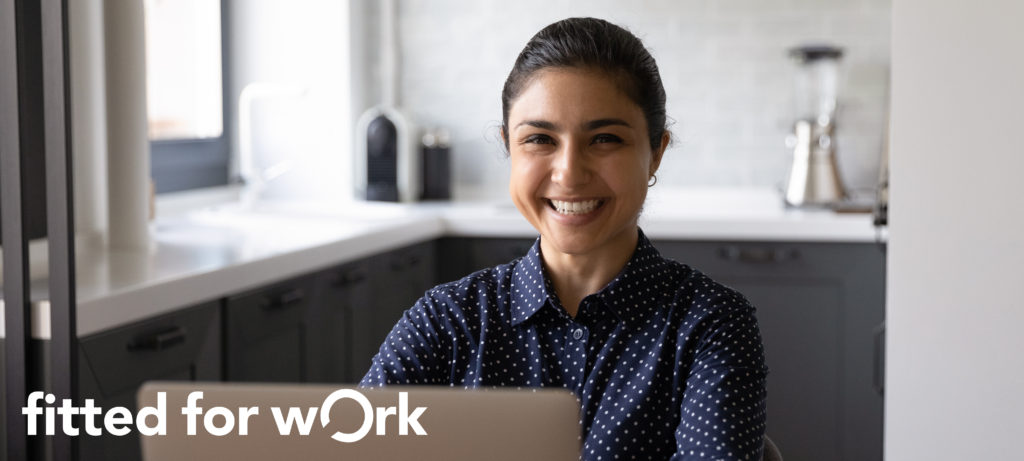 An indian woman wearing a blue shirt with white polka dots is sitting at a computer. She is smiling warmly.