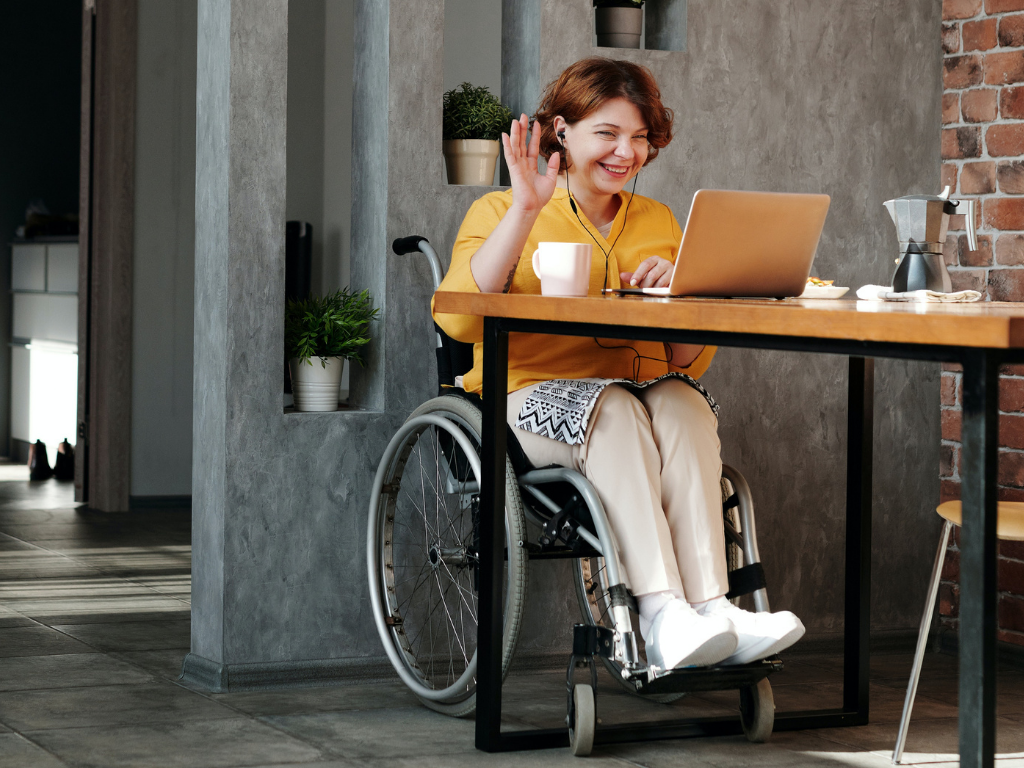 A woman in a wheelchair sits in front of a computer with headphones in. She is in a video meeting and is waving to the other participants.