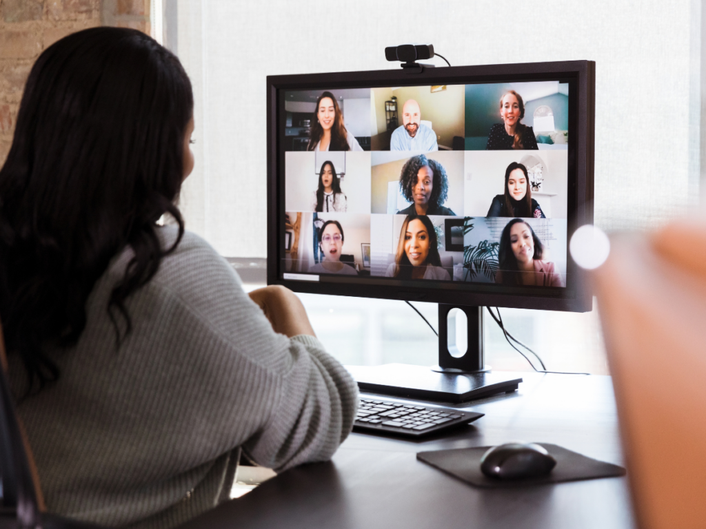 Woman looks at computer. She is in a video call with 9 women and they are smiling at one another.